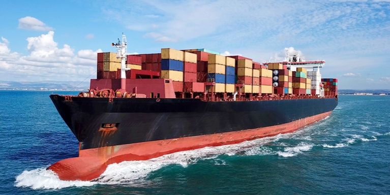 A large cargo ship carrying stacked shipping containers sails through the ocean under a partly cloudy sky.