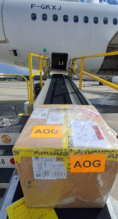 A large cardboard box labeled "AOG" is placed on a conveyor belt leading into the cargo hold of an airplane at an airport.