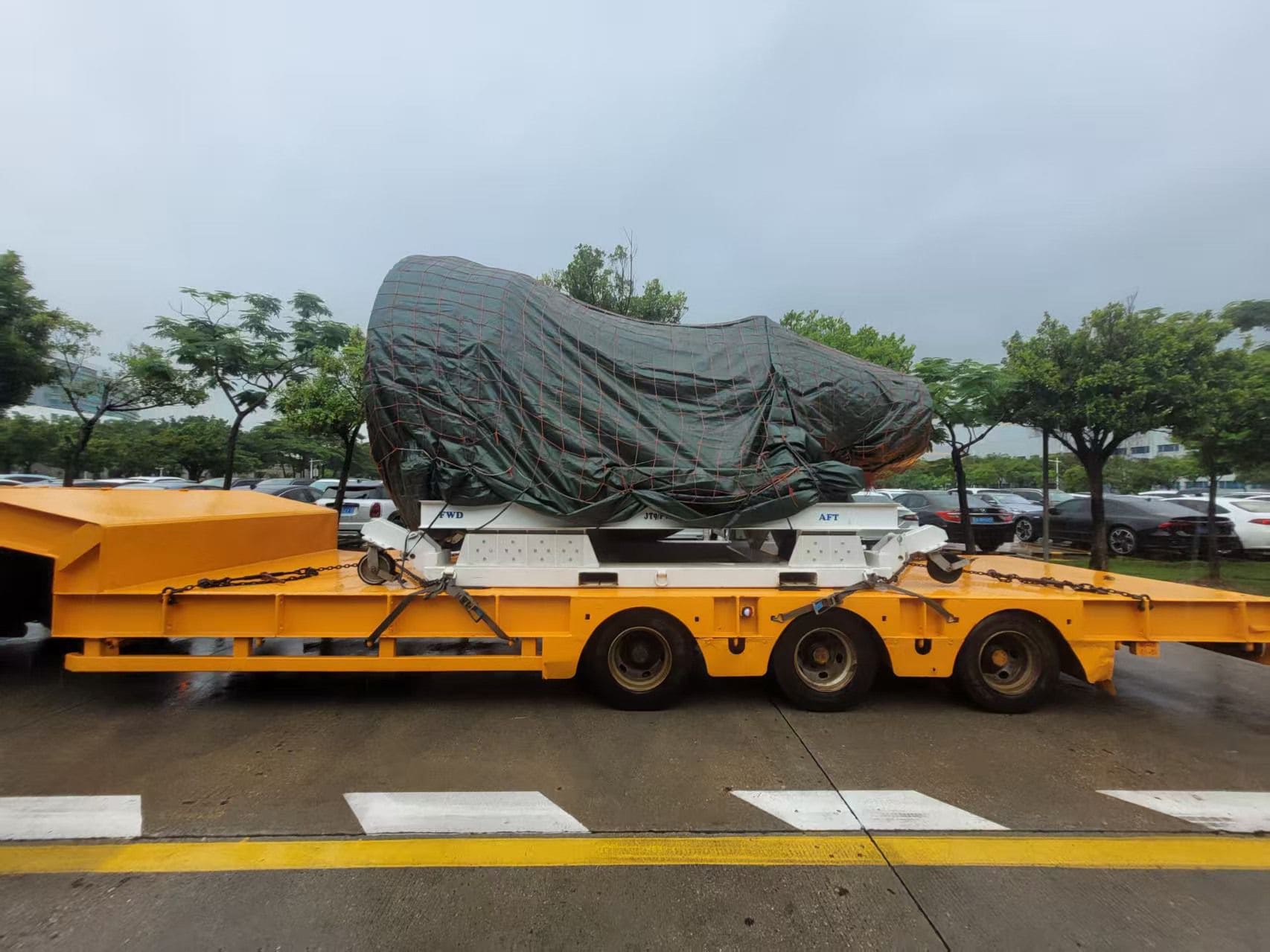 A large object covered with a tarpaulin is secured on a yellow flatbed trailer parked outdoors on a rainy day.