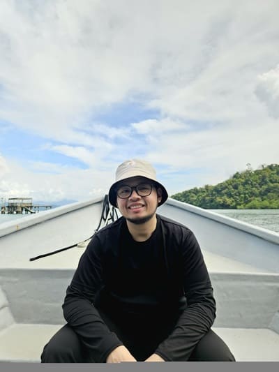 A person wearing a light bucket hat and glasses sits at the front of a boat with water and green hills in the background under a cloudy sky.