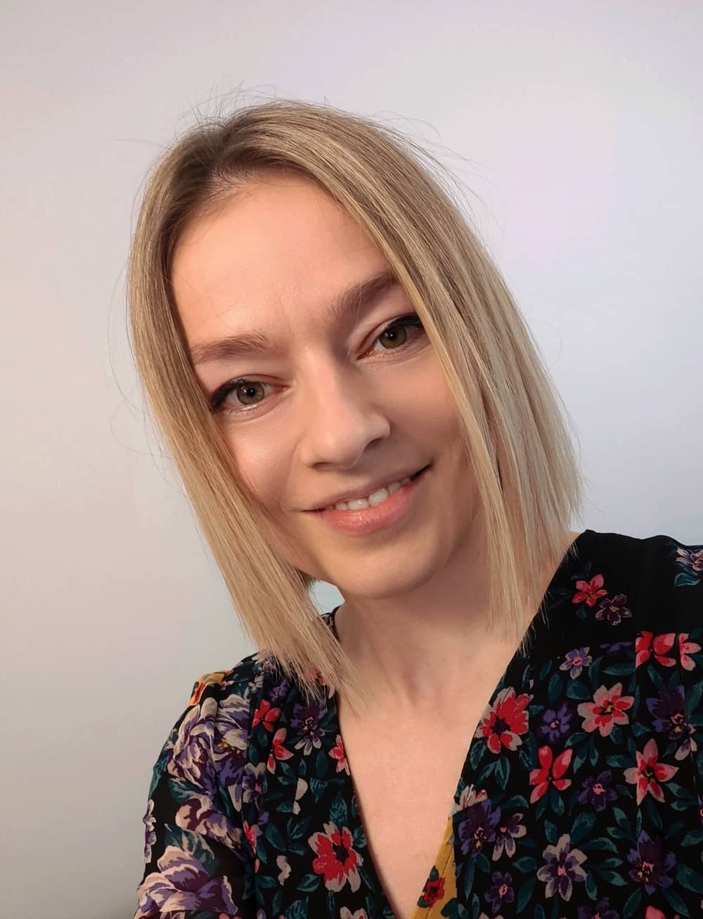 A woman with straight blonde hair and a floral top smiles at the camera in front of a plain light background.