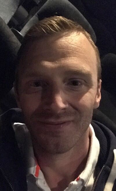 A man with short light brown hair, wearing a white collared shirt and dark jacket, is sitting in a dimly lit room with dark seats visible in the background.