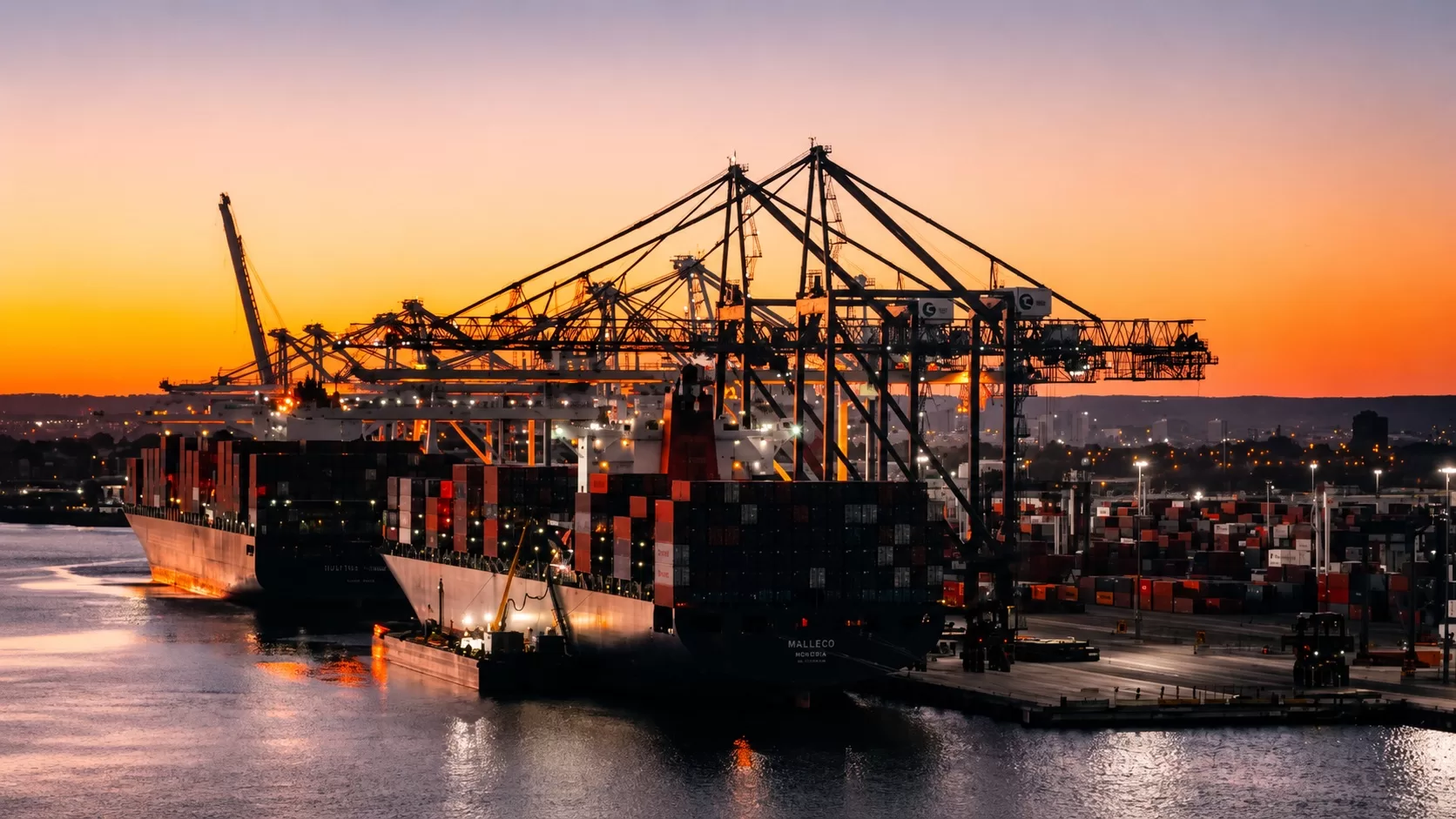Cargo ships docked at a shipping port with large cranes and containers visible at sunset.