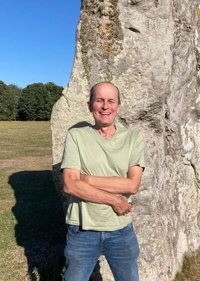 A person stands in front of a large stone outdoors on a sunny day, with arms crossed and trees visible in the background.