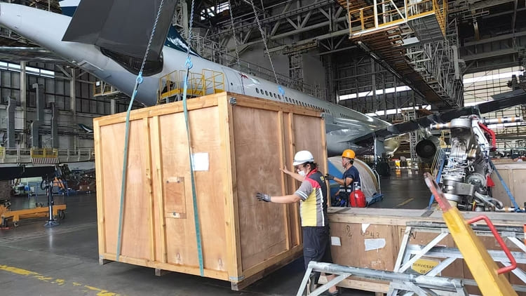 Two workers in a hangar move a large wooden crate near an airplane undergoing maintenance.