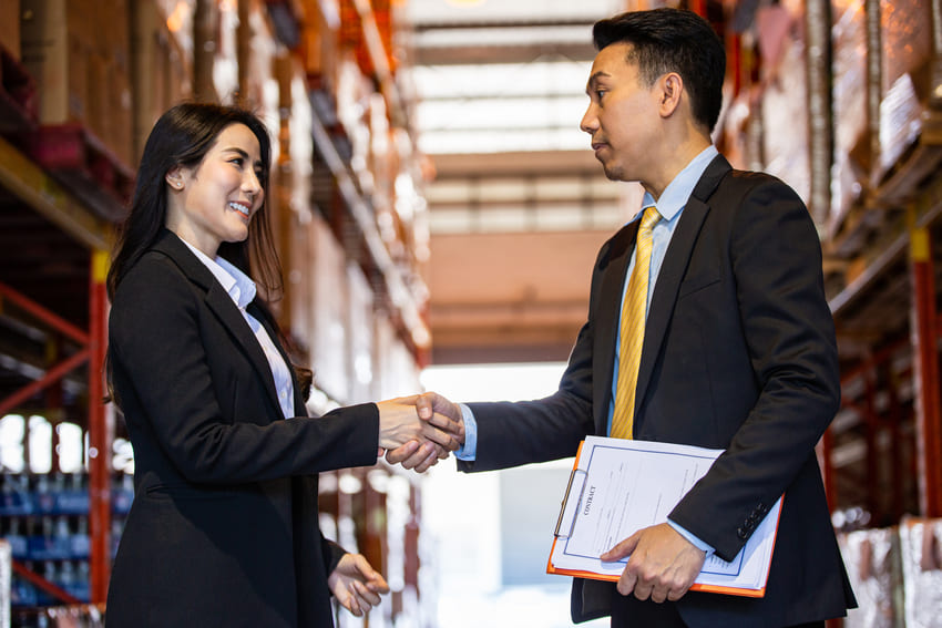 Two business professionals in suits shake hands in a warehouse aisle, with one holding documents and shelves of boxes visible in the background.