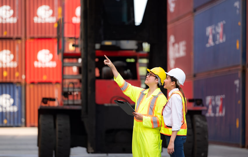 Two workers wearing safety gear stand in front of stacked shipping containers; one holds a clipboard and points while the other watches.