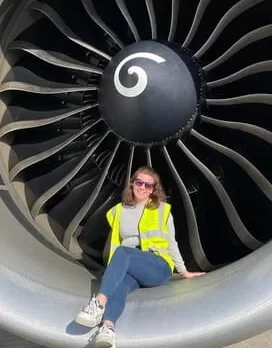 A person wearing a yellow safety vest and sunglasses sits inside the opening of a large jet engine.