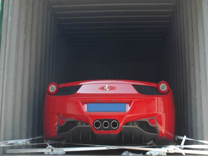 A red Ferrari sports car is parked inside a shipping container, secured with metal braces around the wheels.
