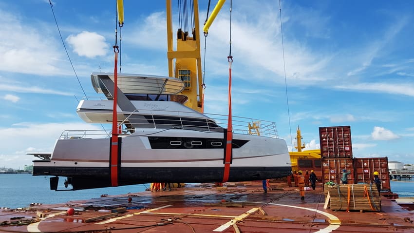 A yacht is being lifted by a crane onto a cargo ship at a port, with shipping containers and workers visible on the deck.