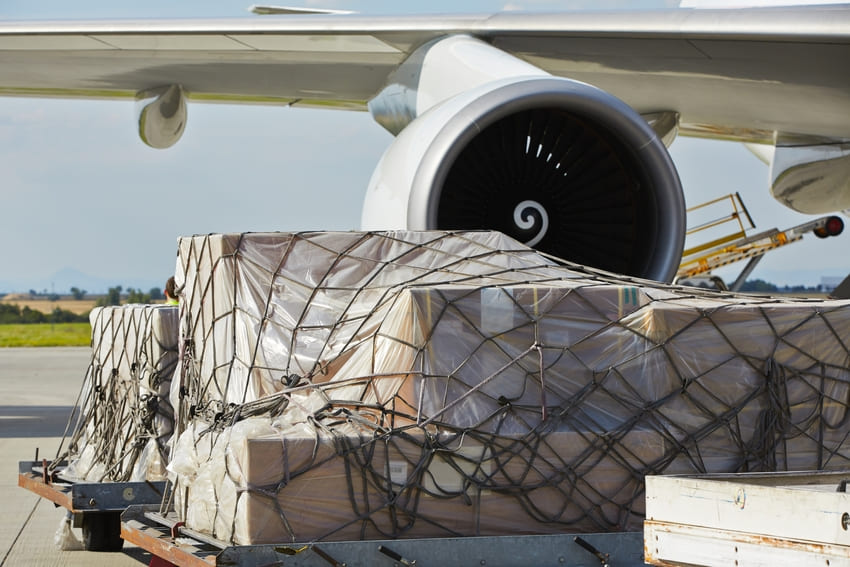 Large cargo pallets wrapped in plastic and netting are loaded onto a jet aircraft on an airport tarmac, with a visible engine in the background.