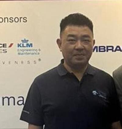 A man in a dark polo shirt stands in front of a white backdrop displaying sponsor logos, including KLM and Embraer.