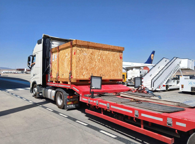 A large wooden crate is loaded onto a red flatbed truck at an airport, with airplanes and mobile stairs visible in the background.
