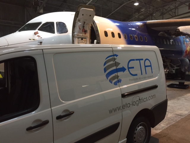 A white ETA Logistics van parked in front of a passenger airplane inside a hangar, with the van’s side door open.