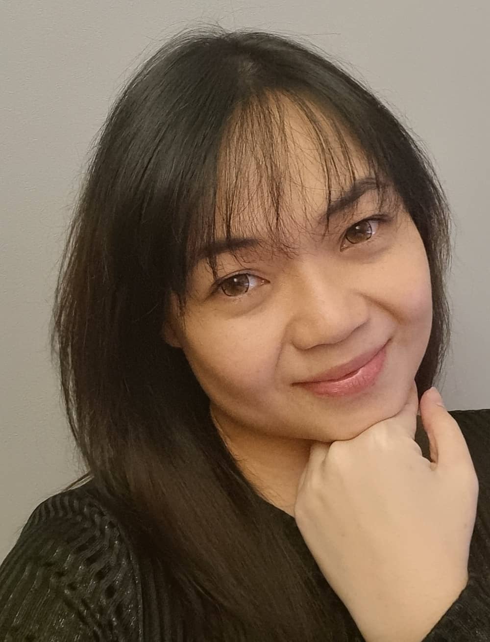 A person with long dark hair and a black top smiles softly at the camera, resting their chin on their hand against a plain light background.