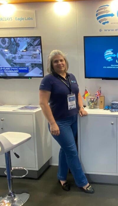 A woman with gray hair stands in front of a booth display wearing a navy polo shirt and jeans, with monitors and flags visible in the background.