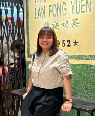 A woman stands and smiles in front of a green and yellow Lan Fong Yuen sign, wearing a light shirt and dark skirt.