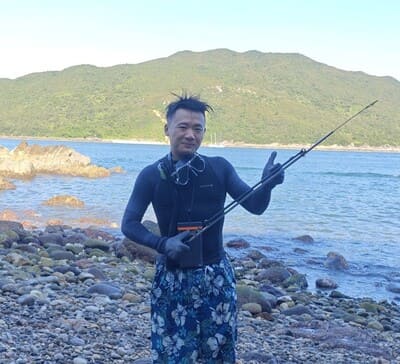 A man in a wetsuit and floral shorts stands on a rocky shoreline holding a fishing rod, with hills and water in the background.