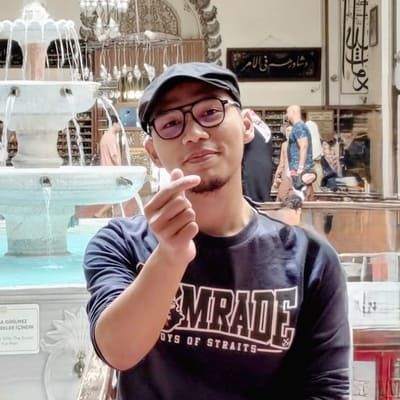 A man wearing glasses and a cap stands in front of a fountain, making a finger heart gesture.