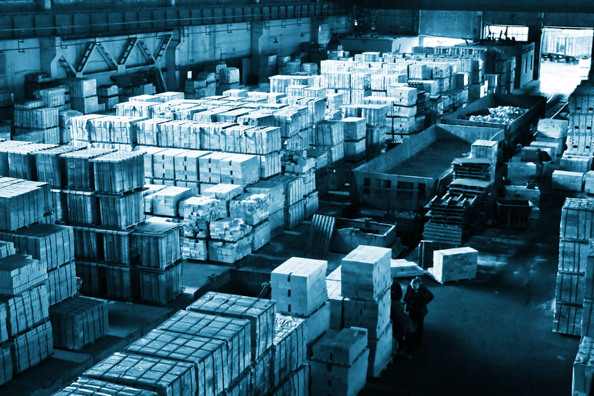 Large warehouse interior filled with neatly stacked pallets and crates, organized in rows under industrial lighting.