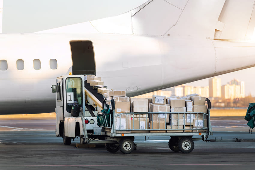A baggage cart loaded with boxes and packages is positioned next to an airplane’s open cargo hold on an airport tarmac.