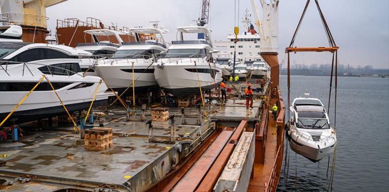 Several yachts are loaded onto a ship’s deck, while a crane lifts another yacht from the water. Several workers in safety gear are present. The background shows a foggy harbor.