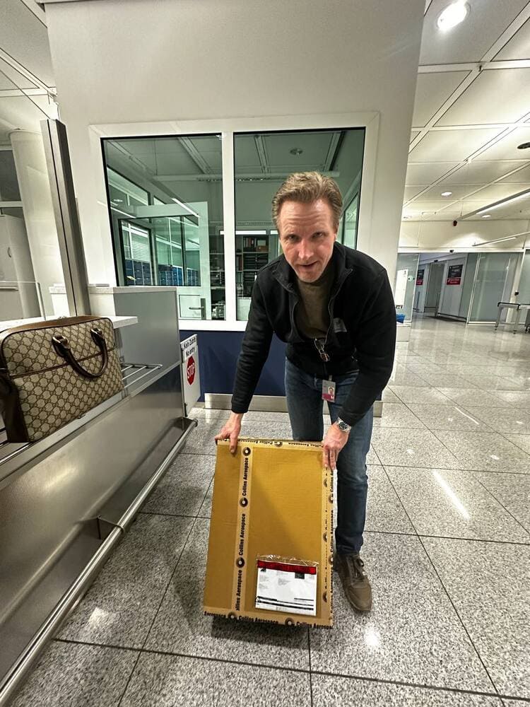 A man stands in an airport security area holding a large, flat, rectangular cardboard package near his feet.
