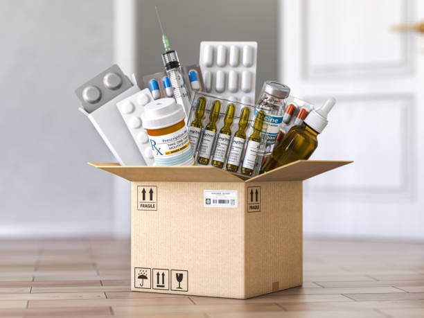 A cardboard box on a wooden floor is filled with various medications, including pill bottles, blister packs, syringes, ampoules, and dropper bottles.