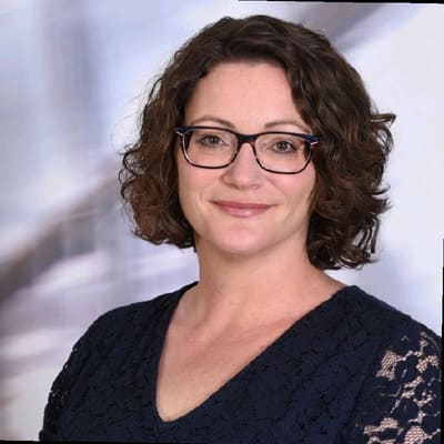 Woman with short curly brown hair and glasses wearing a dark blue lace top, smiling at the camera with a blurred background.