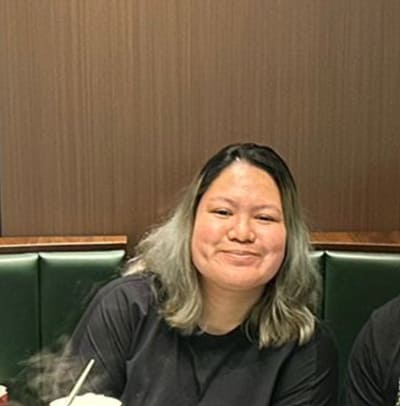 A person with shoulder-length dark hair with lighter ends is sitting at a table in a restaurant, smiling at the camera.