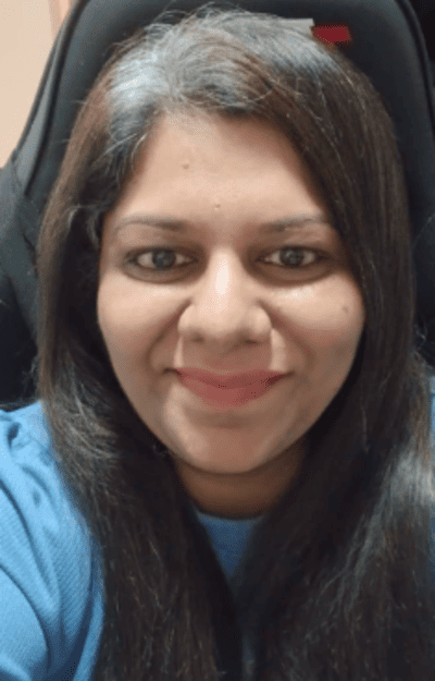 A woman with long, dark hair and a hint of gray is smiling at the camera, wearing a blue top and sitting in a dark chair.