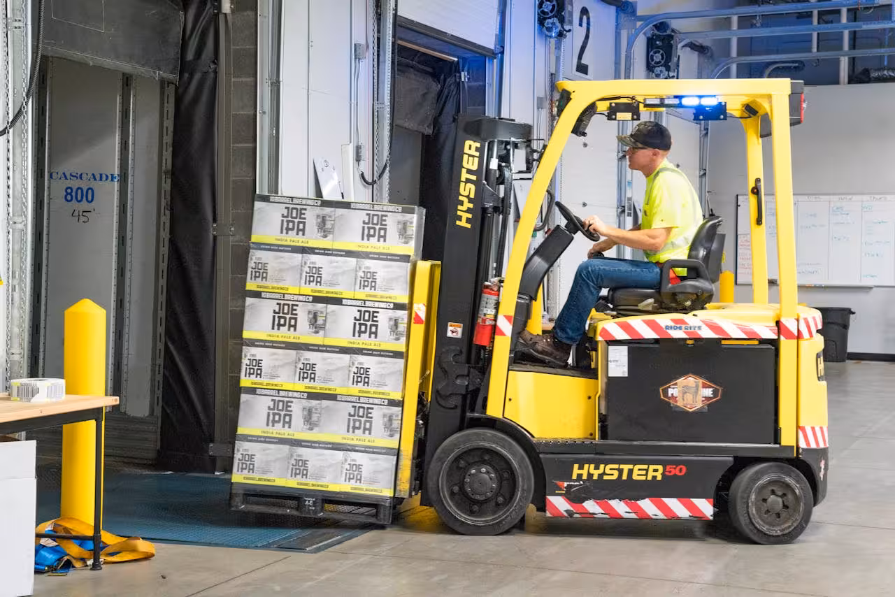 A worker operates a yellow forklift, unloading several boxes labeled "Joe IPA" from the vehicle into a loading dock inside a warehouse.